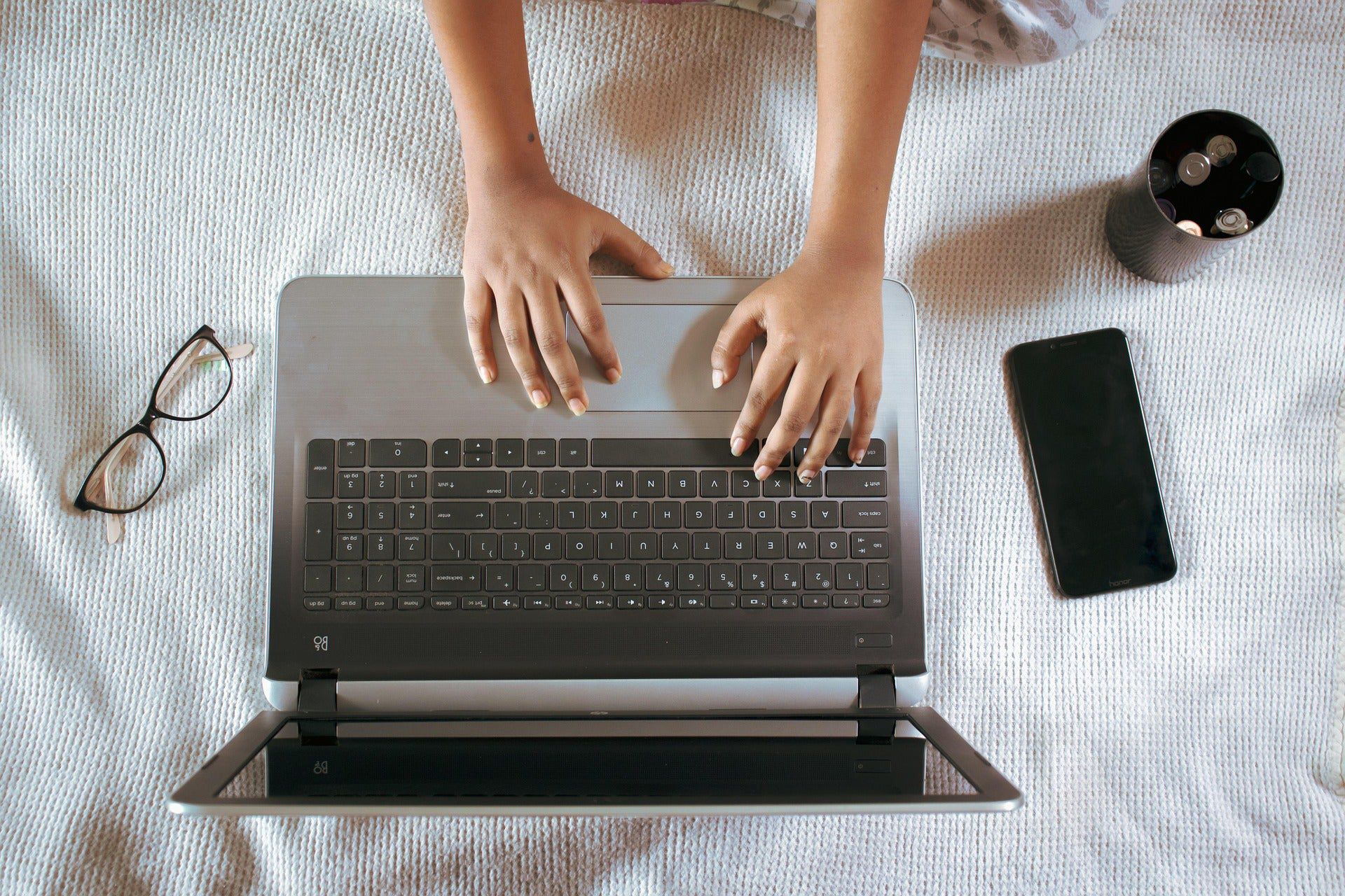 Person using a laptop on a bed with a phone, glasses, and cup nearby